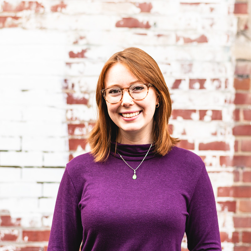 A woman with red hair and a purple sweater, smiling for a portrait outside in front of a brick wall.