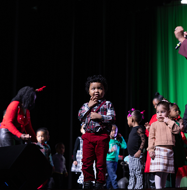 Group of young children standing on stage with a woman in a red sweater, some children wearing festive and plaid outfits, and a green curtain in the background.