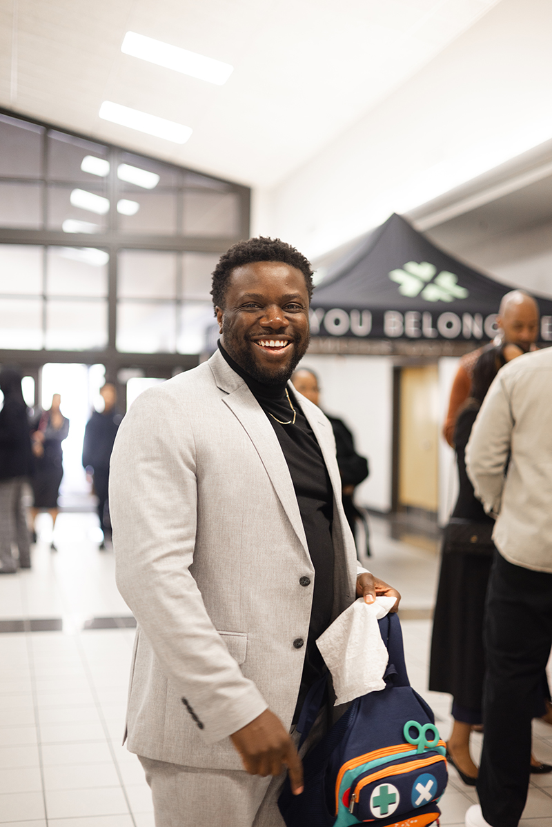 Smiling man in gray suit holding a blue backpack with colorful patches in a bright indoor space.