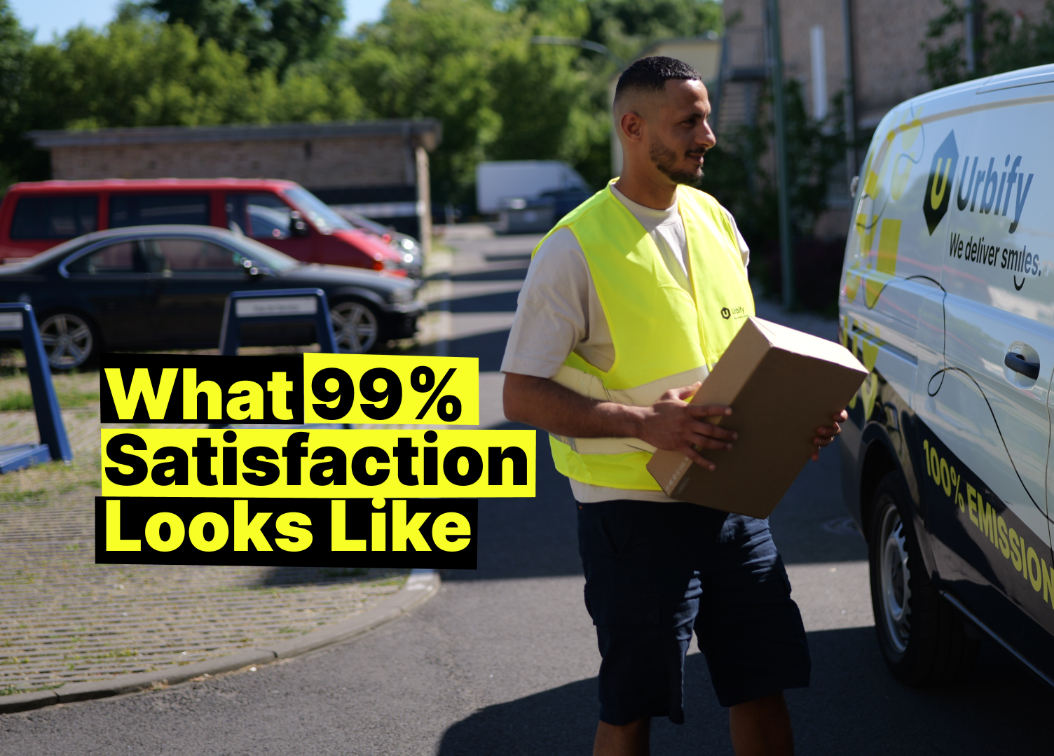 A delivery person in a bright yellow safety vest smiles while holding a cardboard box, standing in front of a white Urbify delivery van with the slogan “We deliver smiles” and “100% EMISSION.” The background features parked cars and greenery on a sunny day