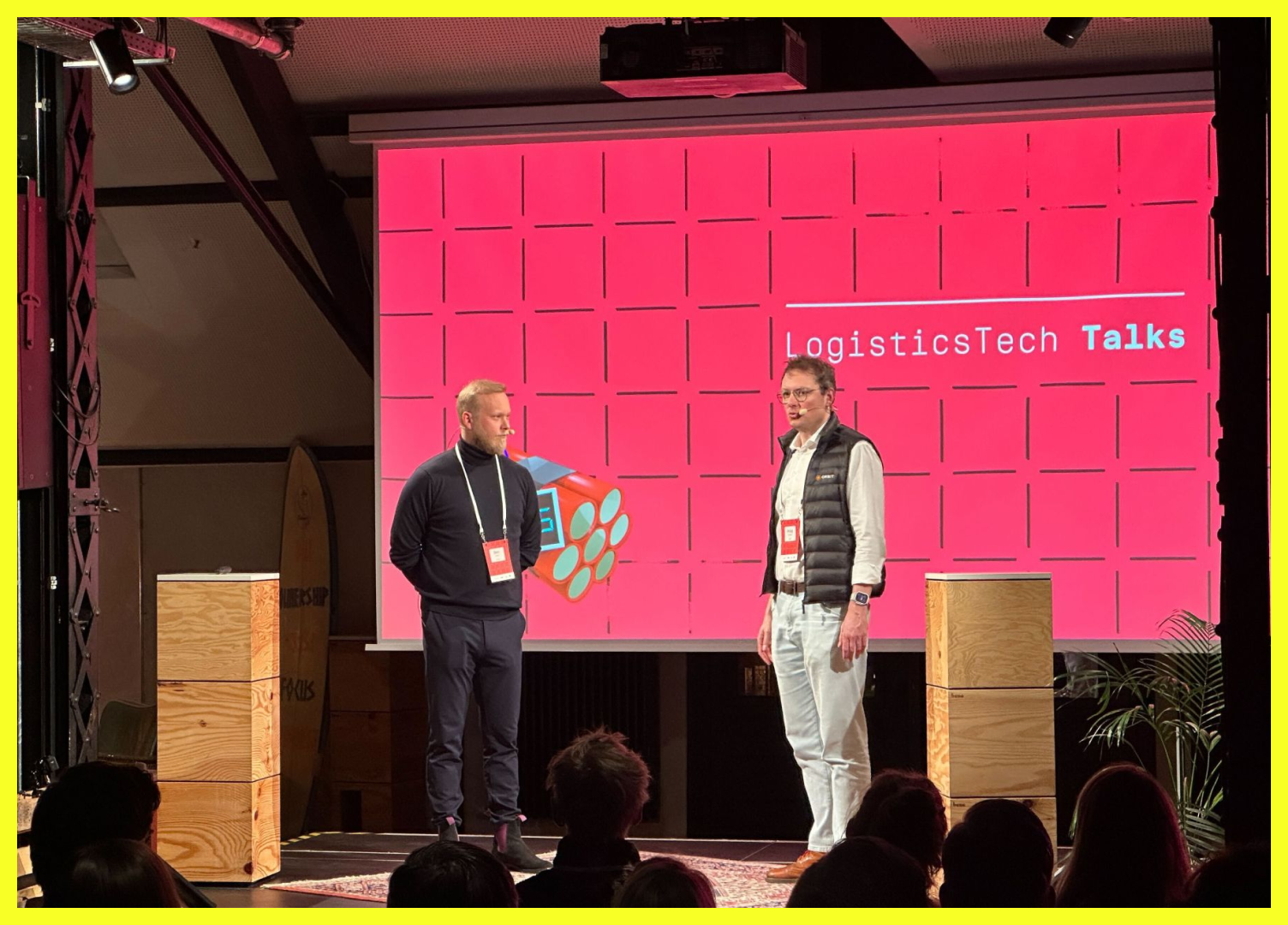 Stage at LogisticsTech Talks with two speakers discussing logistics technology, set against a bright pink backdrop displaying the event name.
