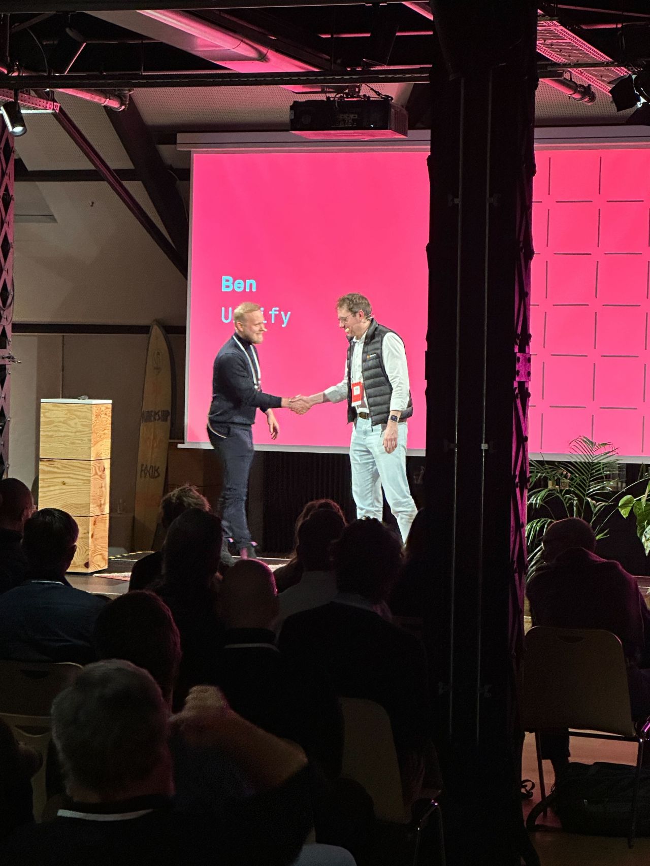 Two men on stage at LogisticsTech Talks, engaged in conversation, with a pink screen and grid pattern in the background.