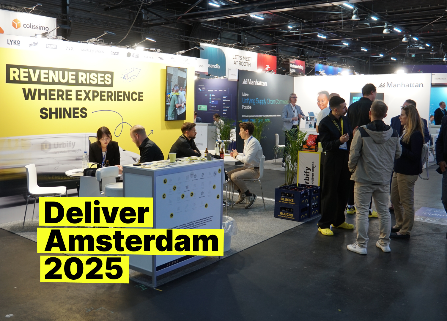 Urbify booth at DELIVER Amsterdam 2025: Team greeting visitors beside a yellow wall reading ‘Revenue rises where experience shines’.