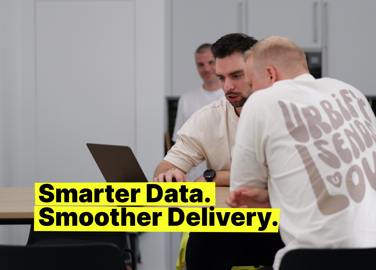 Collaborative team meeting in a modern office: two colleagues discussing delivery data at a laptop, with bold tagline “Smarter Data. Smoother Delivery.” visible.