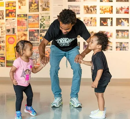 A black man in an art gallery leaning down to hold hands with two very young children