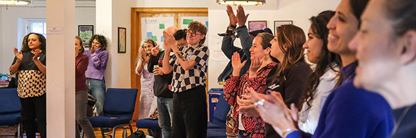 Group of mostly women standing and clapping in a brightly lit meeting room.