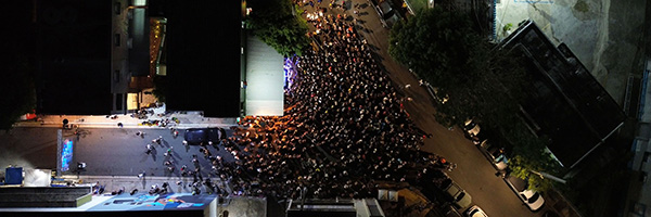 Aerial view of a large nighttime crowd filling a city street around a lit stage.