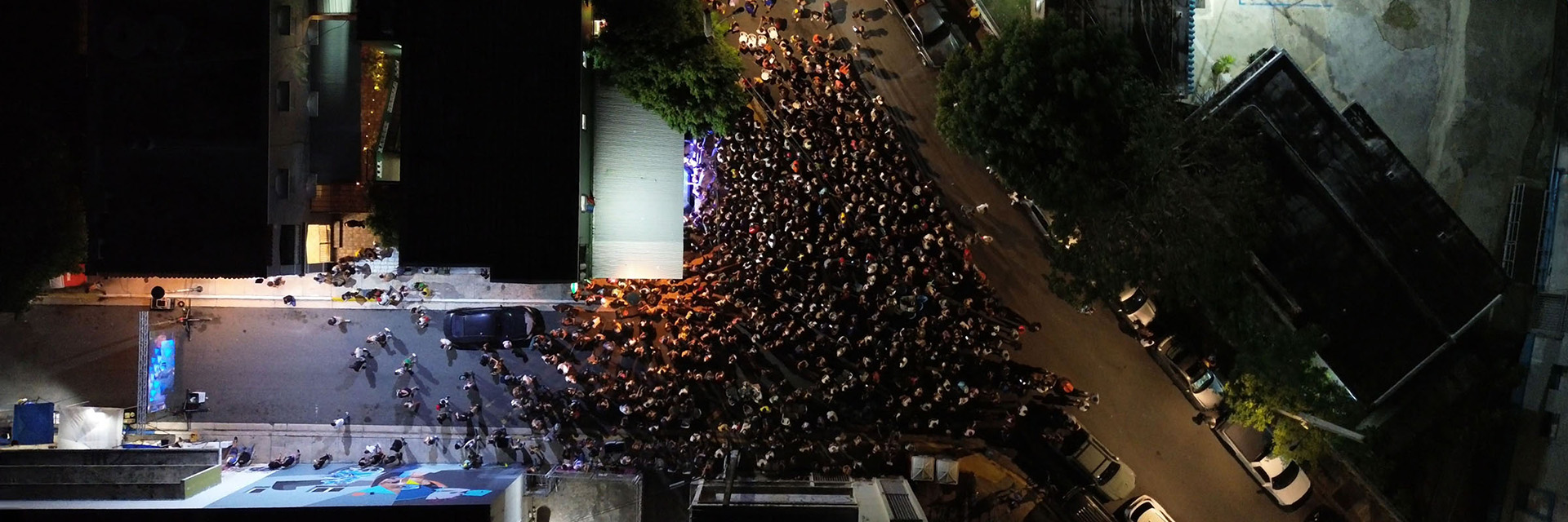 Aerial view of a large nighttime crowd filling a city street around a lit stage.