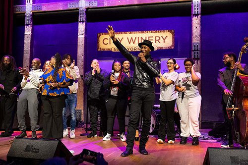 Group of performers onstage at City Winery New York City, with a singer at center raising a hand toward the audience.