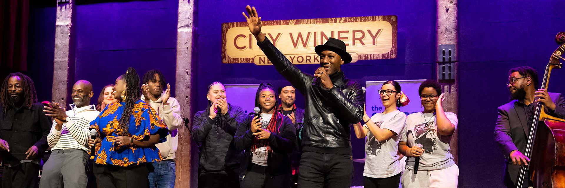 Group of performers onstage at City Winery New York City, with a singer at center raising a hand toward the audience.