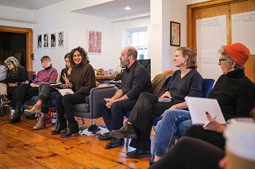 People seated in a circle during a discussion, one person speaking while others listen and take notes.