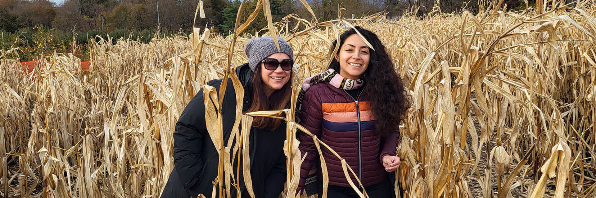 Two women smiling while standing among tall, dried corn stalks on a fall day.