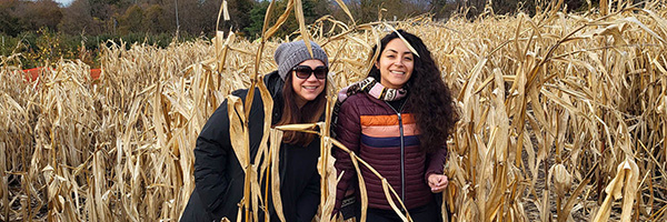 Two women smiling while standing among tall, dried corn stalks on a fall day.