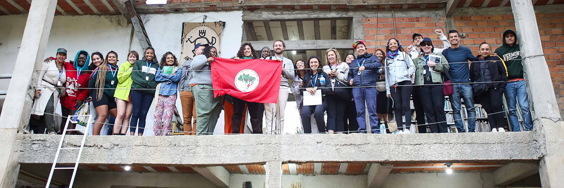 Group standing on a balcony holding the red flag of Brazil’s Landless Workers’ Movement (MST), smiling in solidarity.