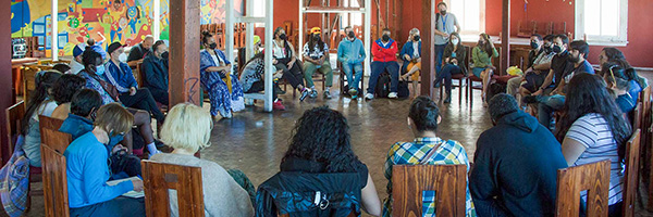 Large group seated in a circle during an indoor community meeting, with colorful murals on the walls.