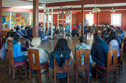 Large group seated in a circle during an indoor community meeting, with colorful murals on the walls.
