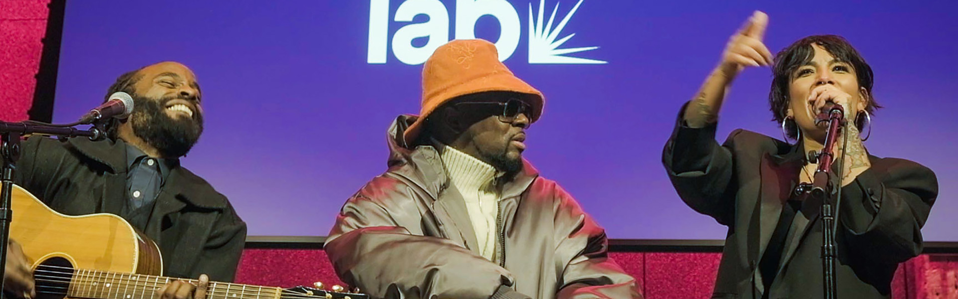 Three musicians performing onstage, one singing into a microphone beside a guitarist and another wearing an orange hat.