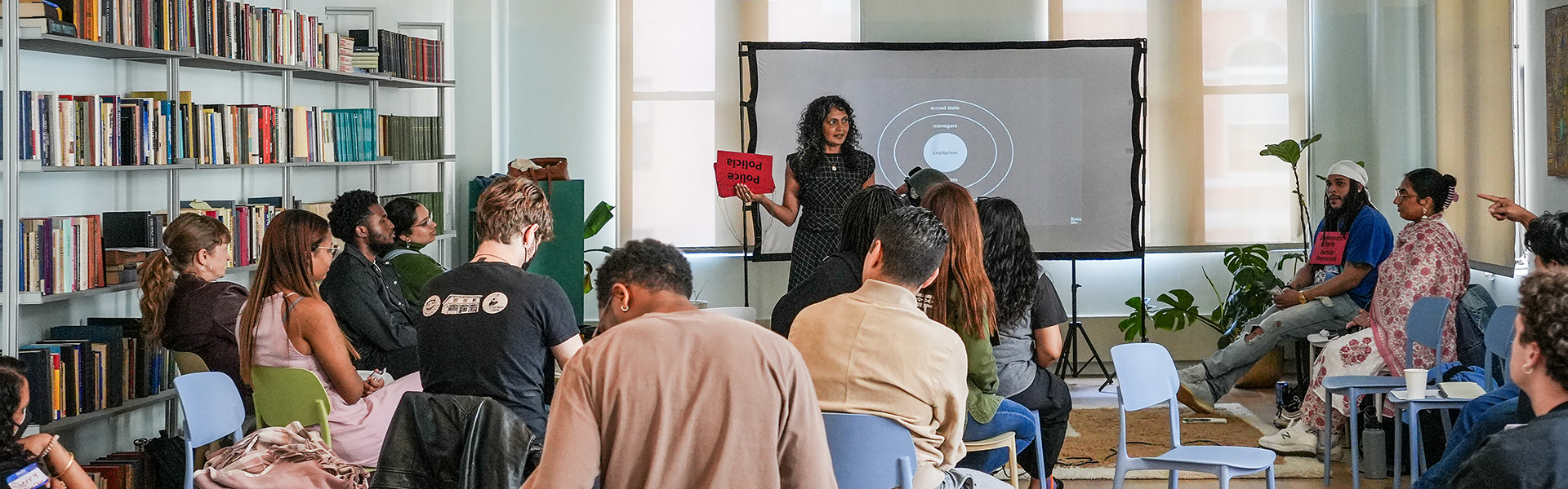 Group of people listening to a presenter holding a red sign in front of a projector screen.