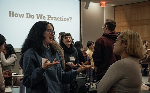 People engaged in animated discussion during a workshop with a slide reading “How Do We Practice?” in the background.