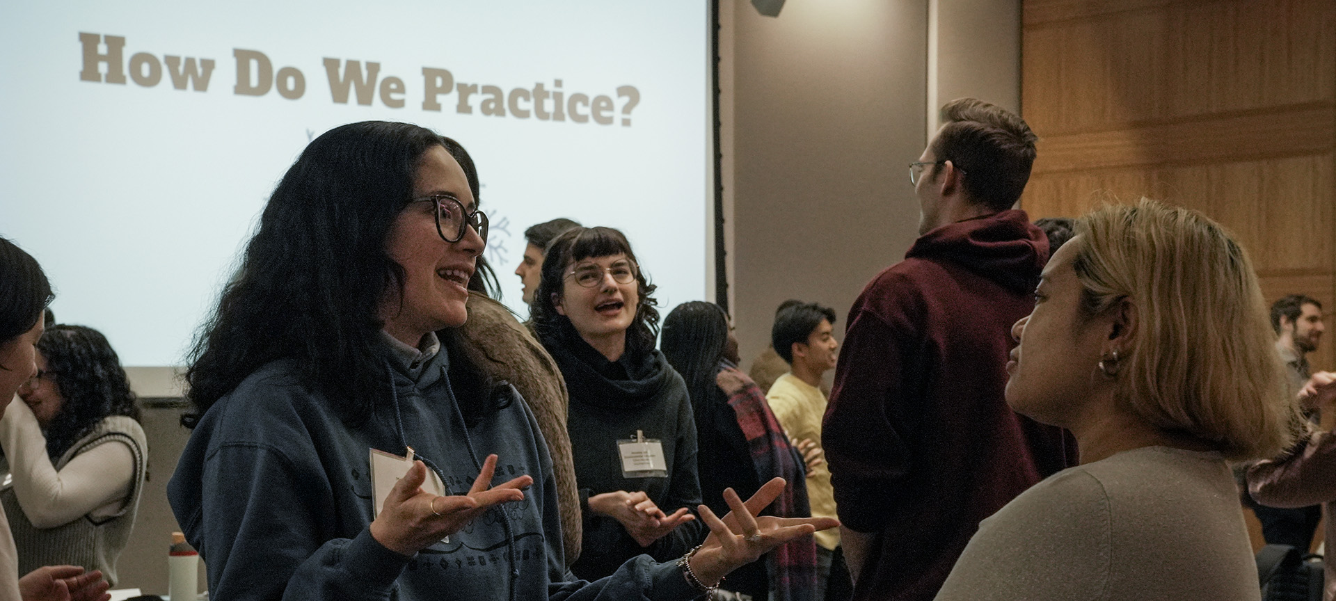 People engaged in animated discussion during a workshop with a slide reading “How Do We Practice?” in the background.