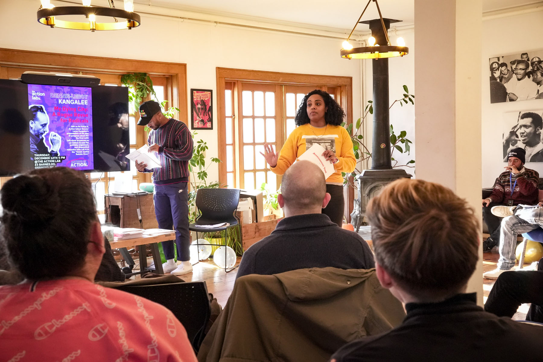 Presenter addressing a seated group in a sunlit room with a screen displaying event details