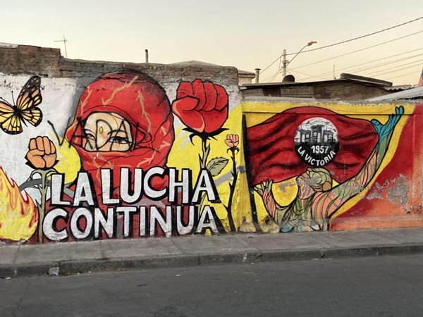 Colorful mural reads “La Lucha Continua” with raised fists, a masked figure, and movement imagery.