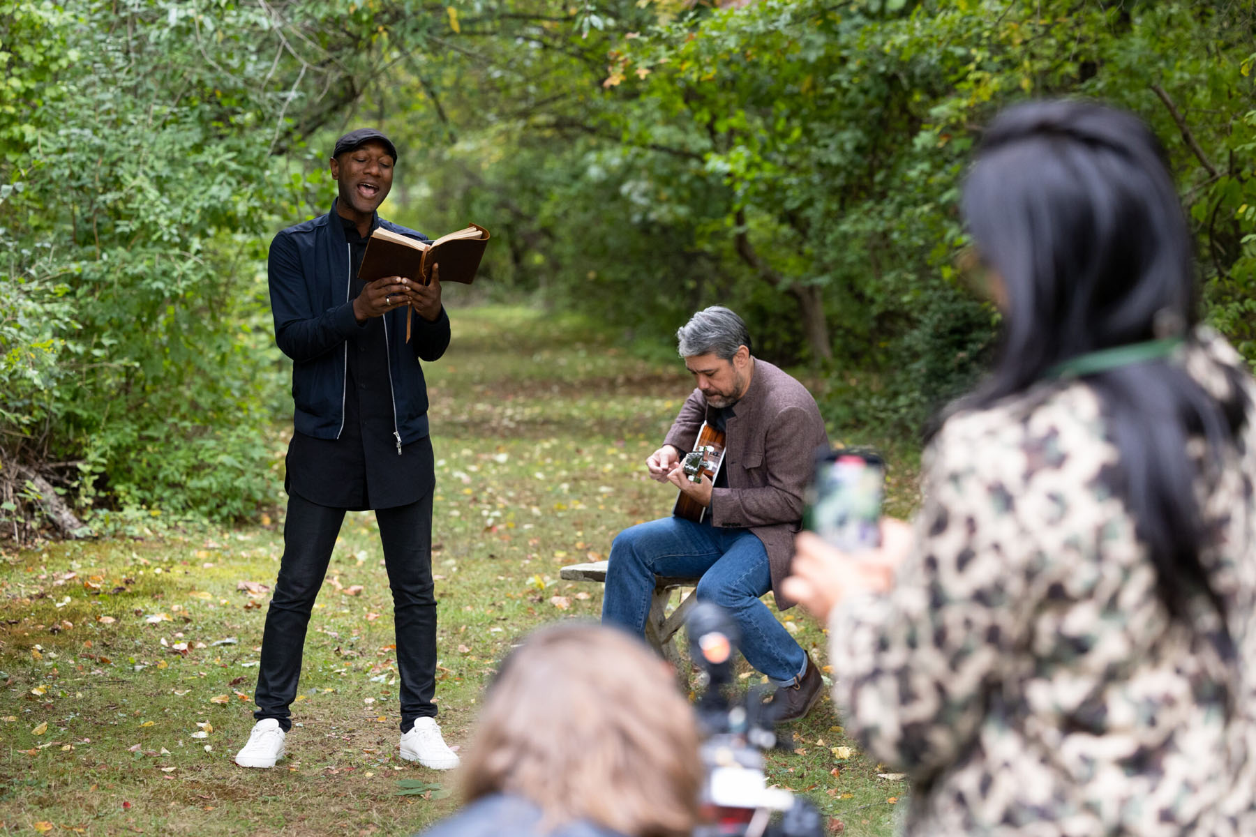 Performer reads from a book as a guitarist plays in a wooded outdoor path.