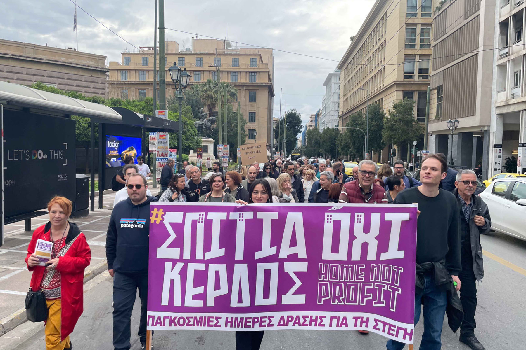 Marchers holding a large purple banner reading “Homes Not Profit” during a street protest