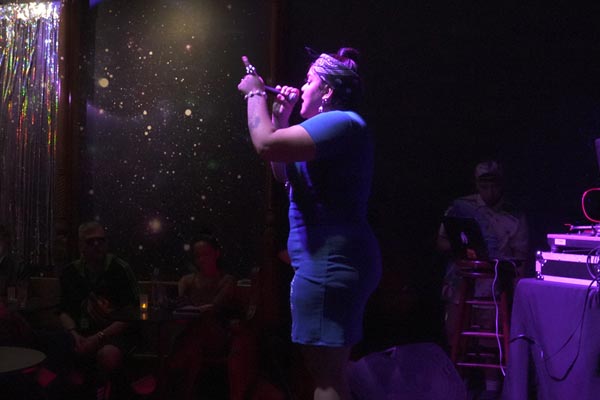 Performer in a blue dress sings on a dimly lit stage before an audience.