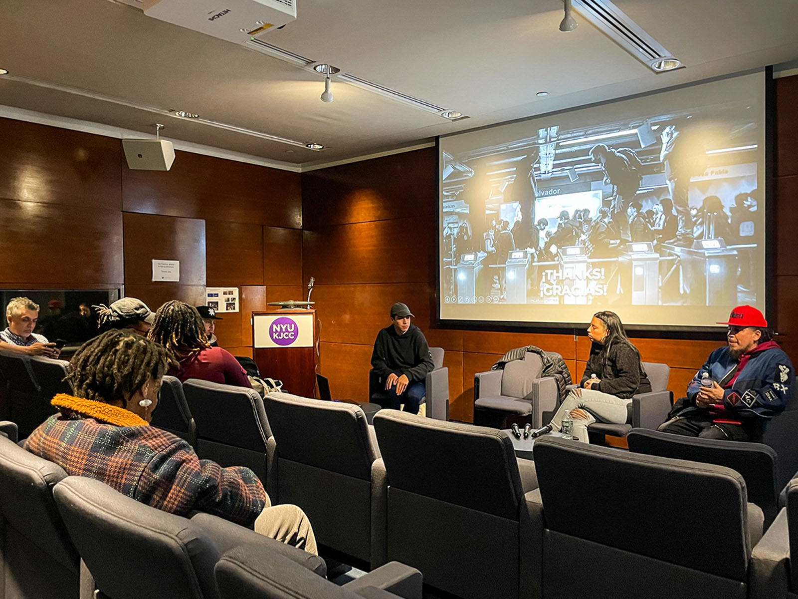 Small audience listens to three speakers seated beneath a large projected black-and-white image.
