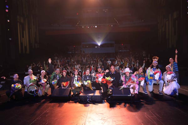 Performers posing onstage with flowers before a cheering audience in a large theater