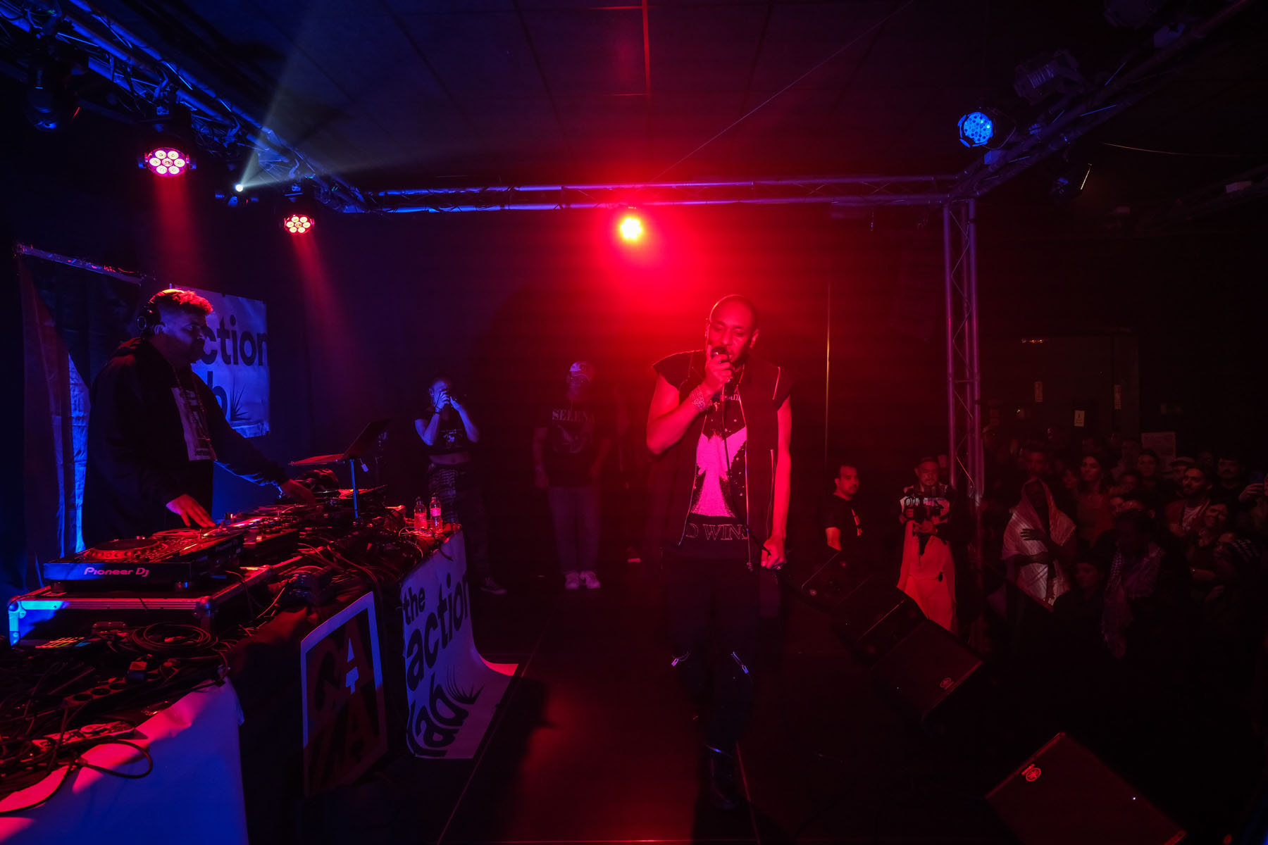 Rapper performing under red stage lights as a DJ and crowd look on