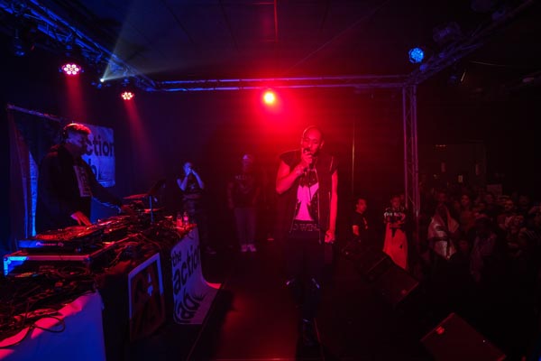 Rapper performing under red stage lights as a DJ and crowd look on