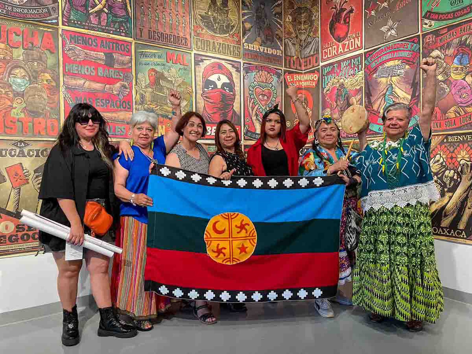 Group of women hold a Mapuche flag in front of a wall of vibrant political posters.