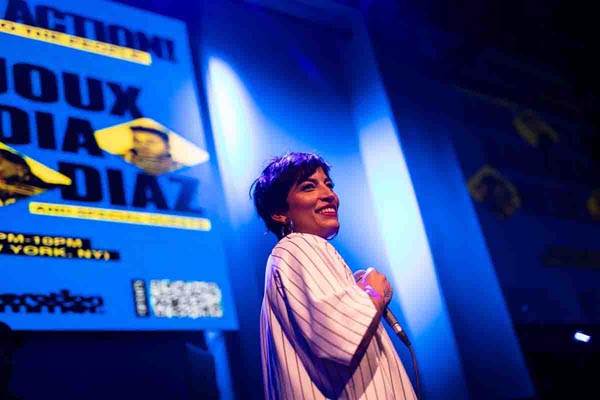 Performer smiles onstage under blue lights with a large event poster behind her.