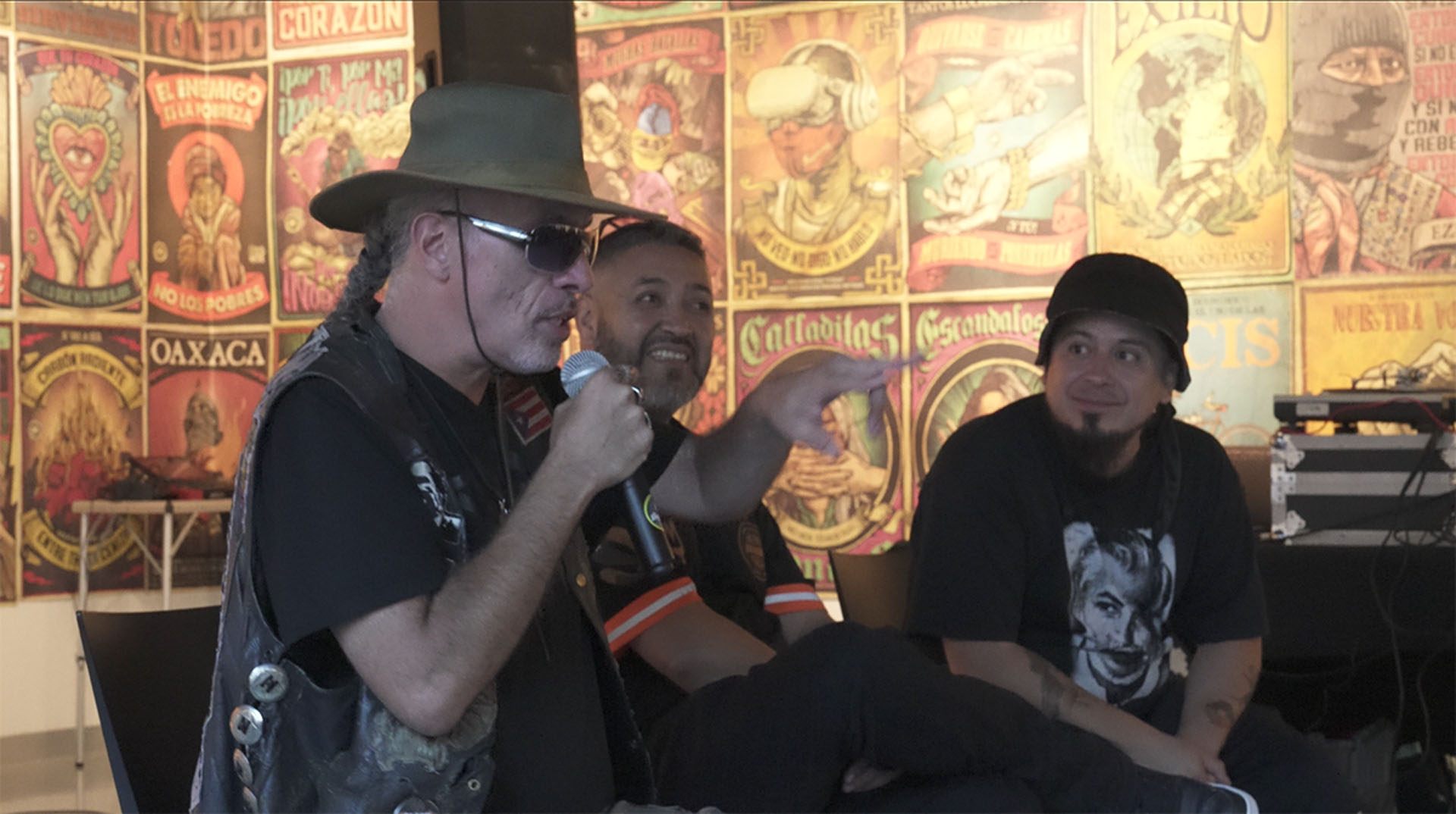 Man in a hat speaks into a microphone as two others listen before a wall of colorful posters.