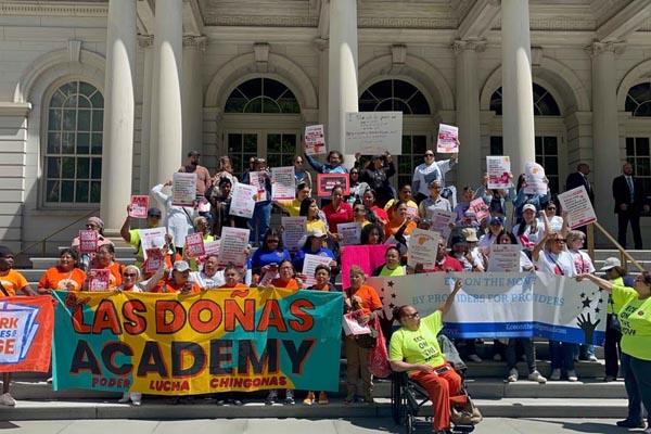 Group gathered on government building steps holding signs and a “Las Doñas Academy” banner