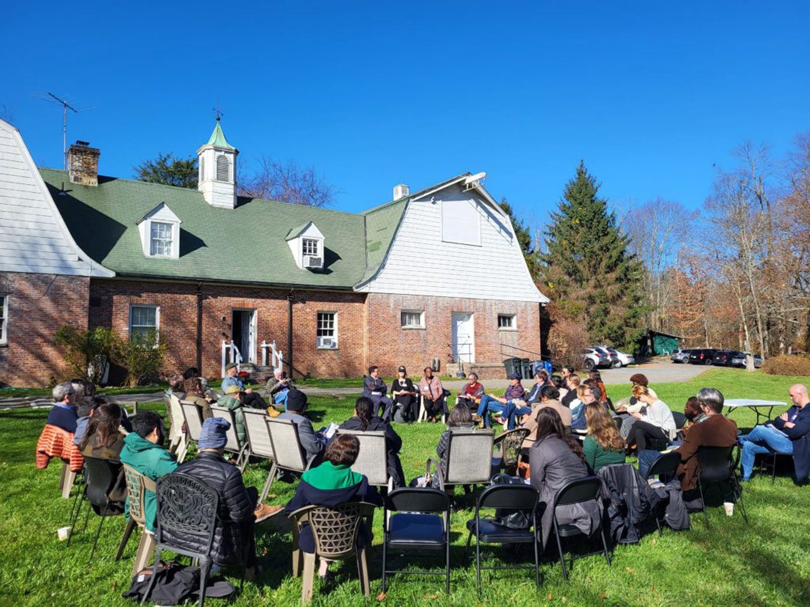 Large group sits in a circle on a sunny lawn outside a historic brick building.