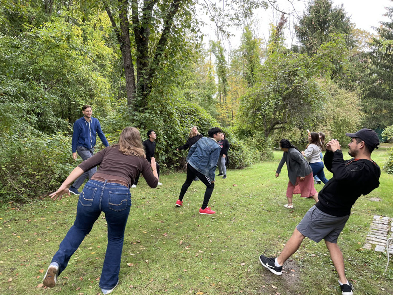 Outdoor photo of a group of people laughing and leaning while playing an activity on a grassy path surrounded by trees.