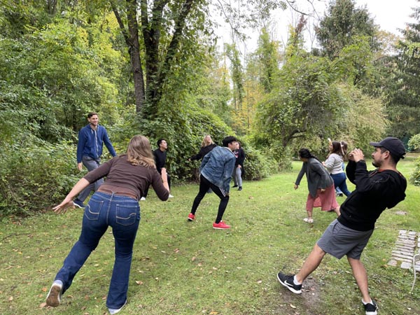 Outdoor photo of a group of people laughing and leaning while playing an activity on a grassy path surrounded by trees.