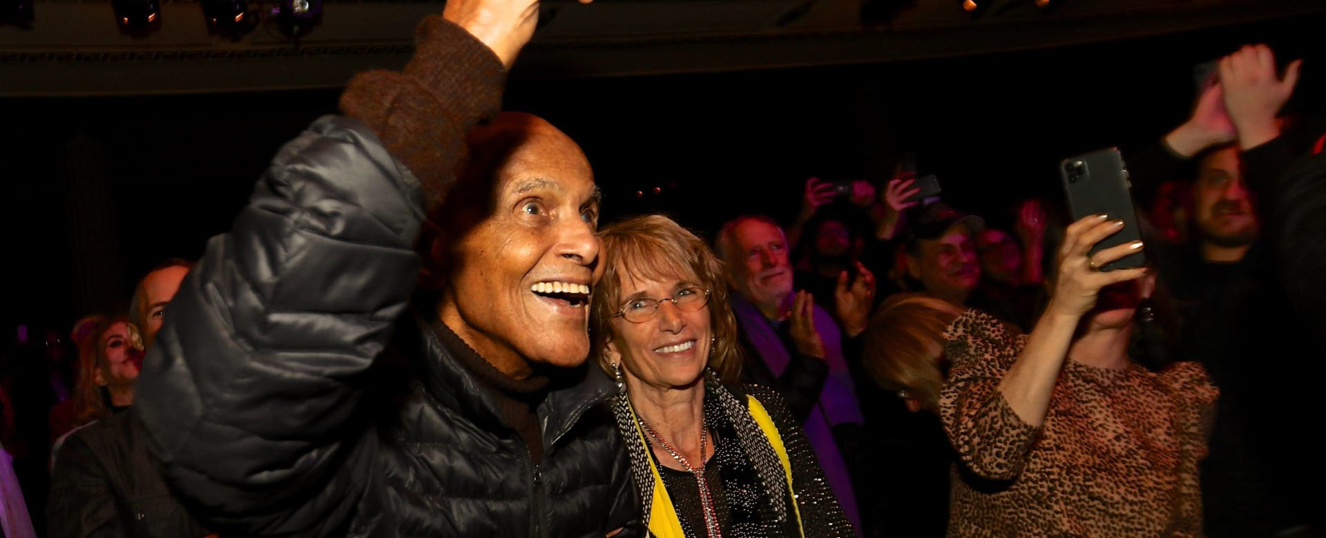 Audience photo showing two people smiling and cheering at a live event, with a crowd behind them taking photos.