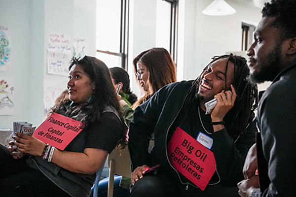 Participants in a workshop laughing and interacting, wearing red signs labeled “Finance Capital” and “Big Oil.”