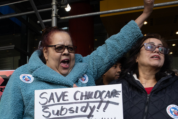 Two women at a rally holding a sign that says “Save Childcare Subsidy!!” while raising a hand and shouting.