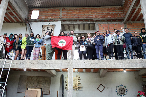Group standing on a balcony holding the red flag of Brazil’s Landless Workers’ Movement (MST), smiling in solidarity.