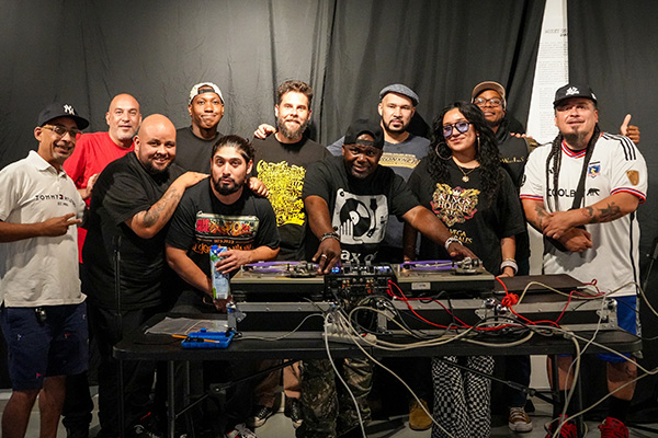Group of DJs and musicians posing together behind turntables and audio equipment.