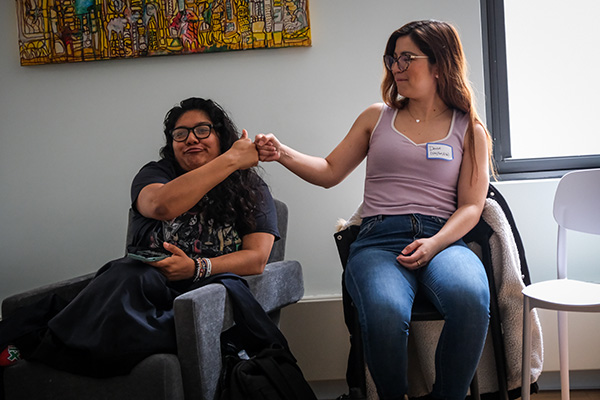 Two young women seated indoors smiling and bumping fists in a friendly gesture.