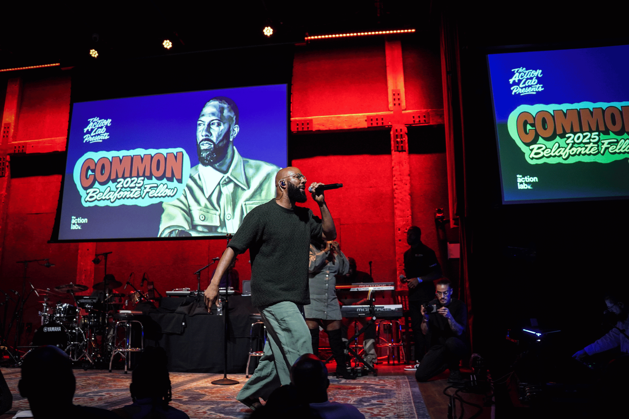 Common with a mic performing in front of a lively audience with red backdrop.