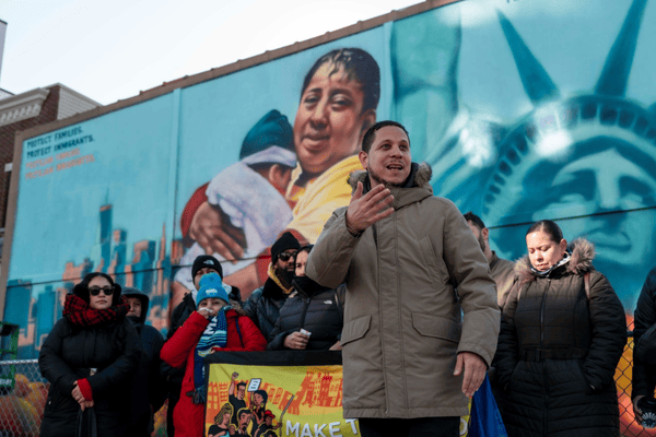 Crowd gathers behind speaker in beige/green parka speaking before a pro immigrant mural with image of statue of liberty poking out.