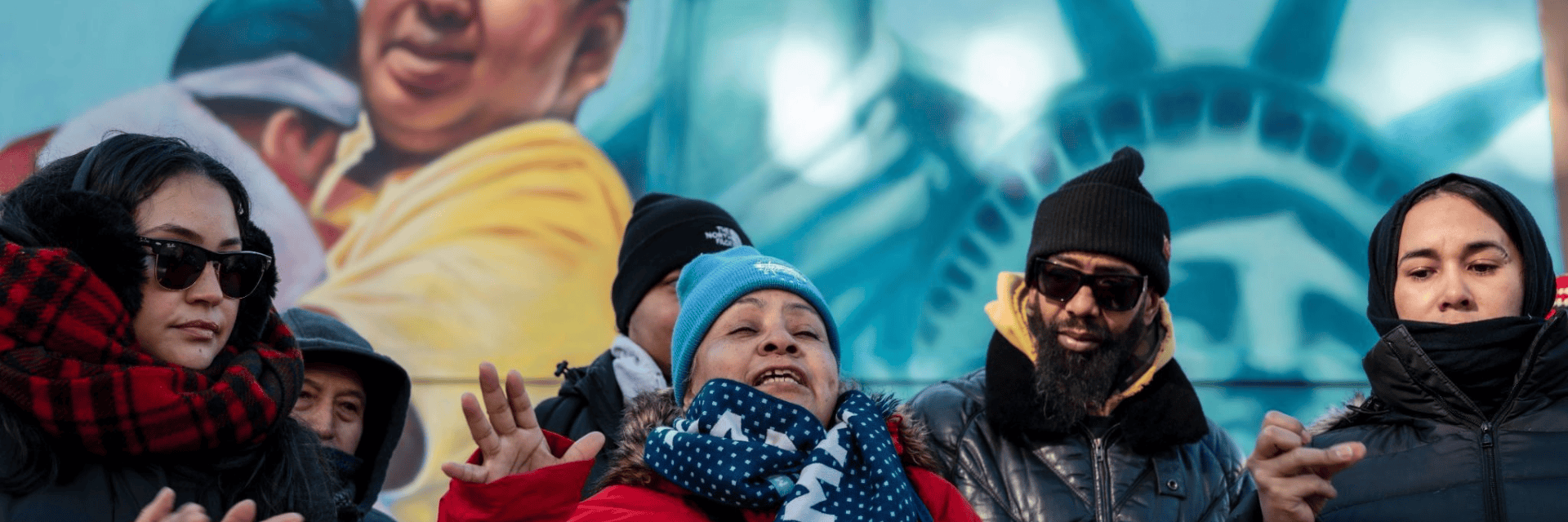 Crowd gathers behind speaker in beige/green parka speaking before a pro immigrant mural with image of statue of liberty poking out.
