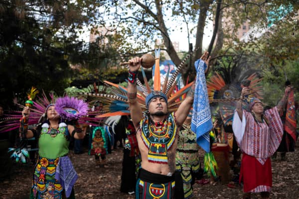 Group performs ceremonial dance in colorful feathered regalia, arms raised outdoors.
